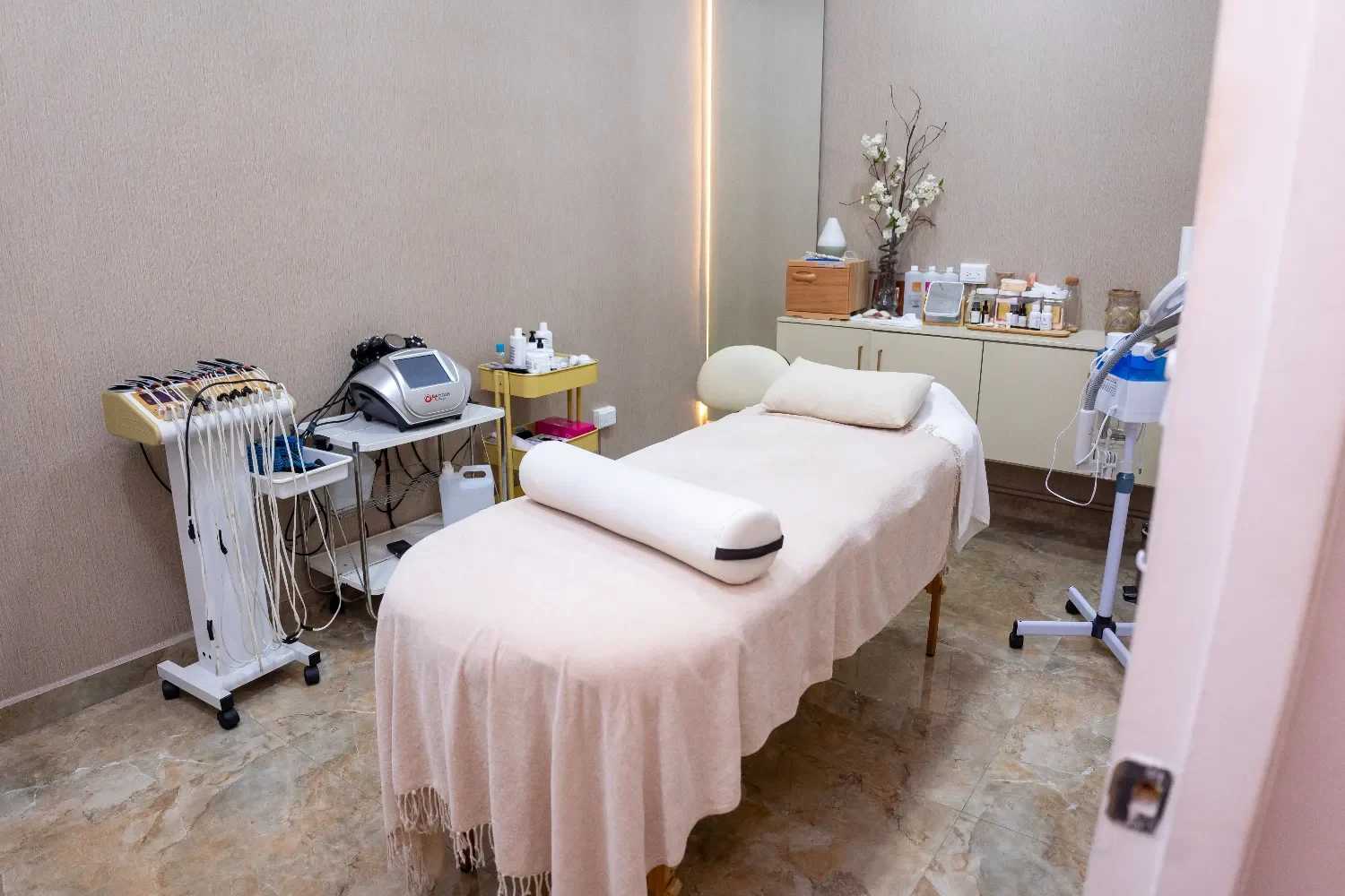 Una sala de tratamiento profesional bien iluminada con una camilla de masaje de color beige cubierta con una manta rosa claro, rodeada de varios equipos de belleza y fisioterapia, paredes texturizadas en tonos beige y suelos de mármol.